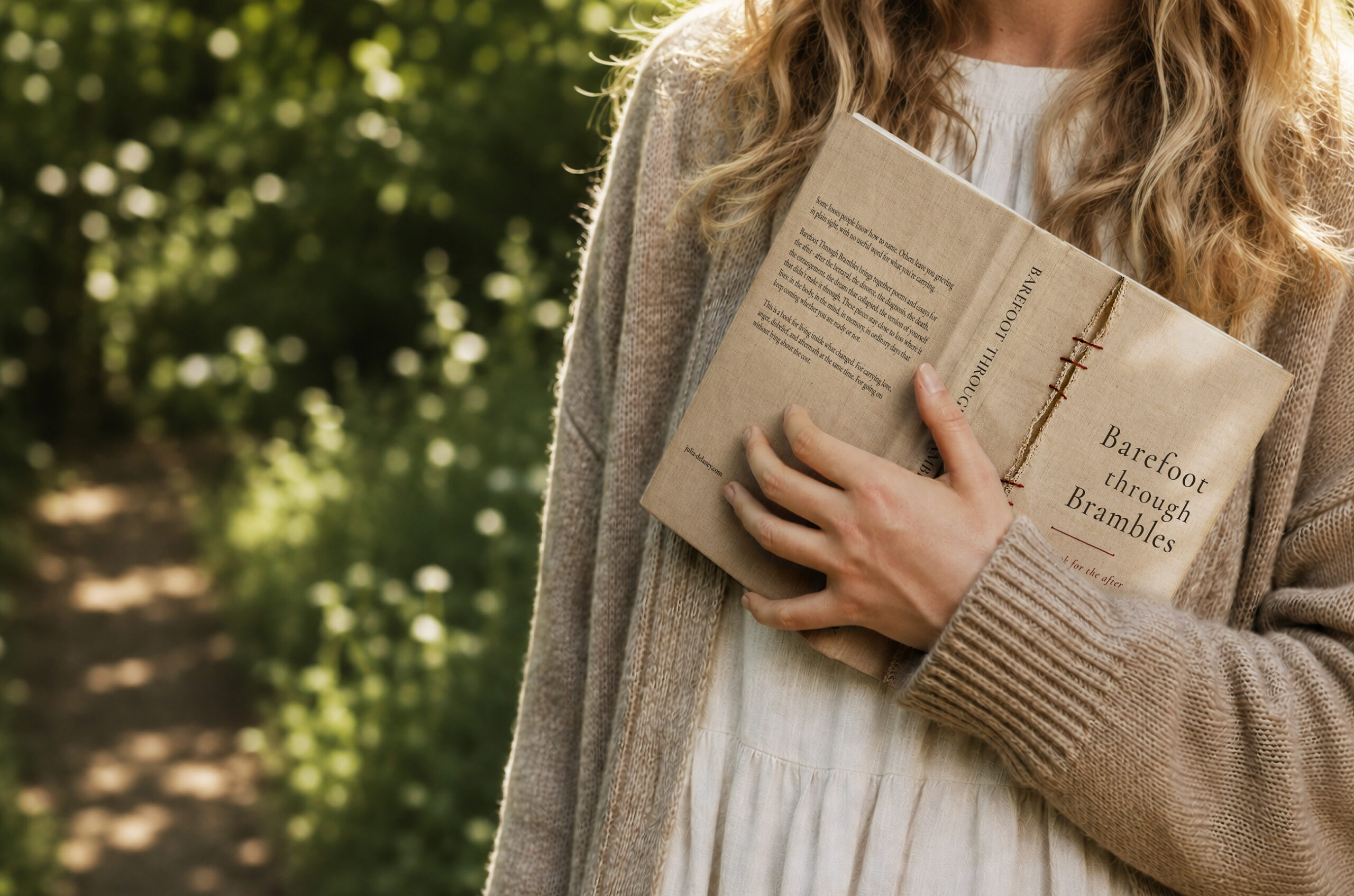 A woman in a cream dress and soft cardigan holds the hardcover edition of Barefoot Through Brambles by Julia Delaney on a sunlit woodland path.