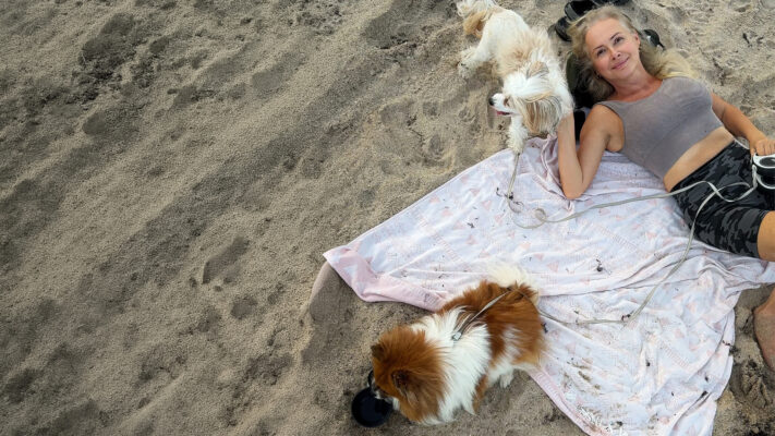 Julia Delaney relaxing on a beach blanket with two small dogs beside her on the sand.