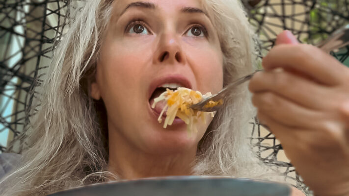 Julia Delaney seated outdoors with a bowl of noodles, lifting a forkful to eat in a casual everyday moment.