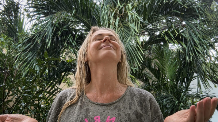 Julia Delaney standing beneath palm trees in heavy rain with wet hair, eyes closed, and her face lifted toward the rainfall.